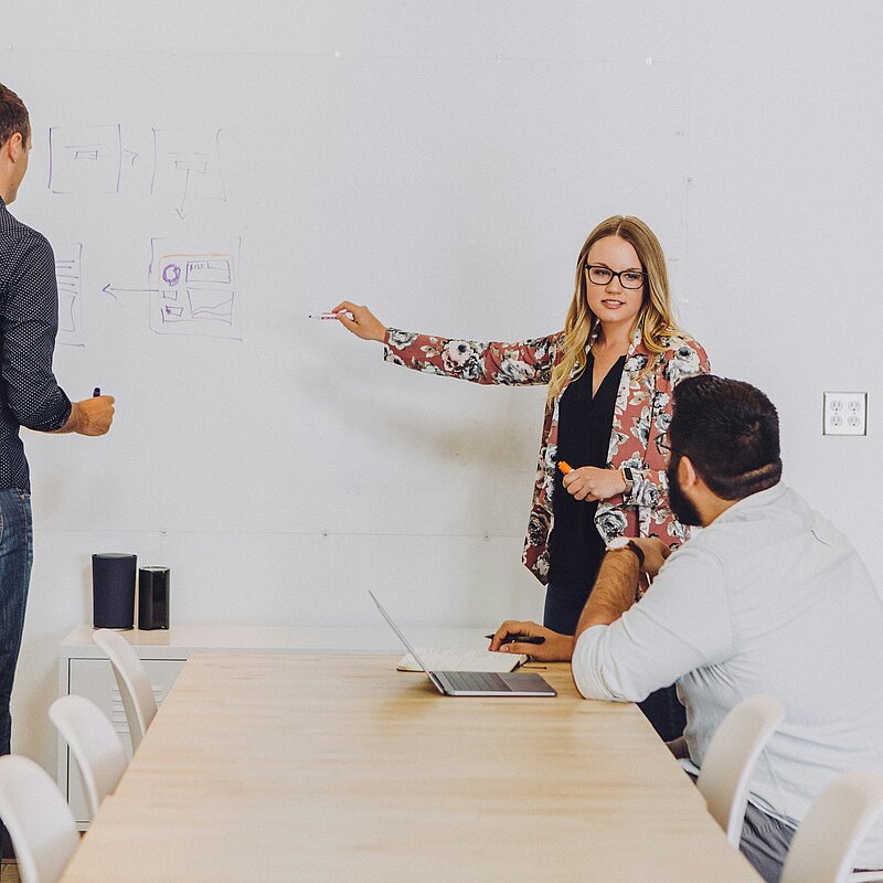 Frau zeigt ein Diagramm am Whiteboard, zwei Männer schauen zu.