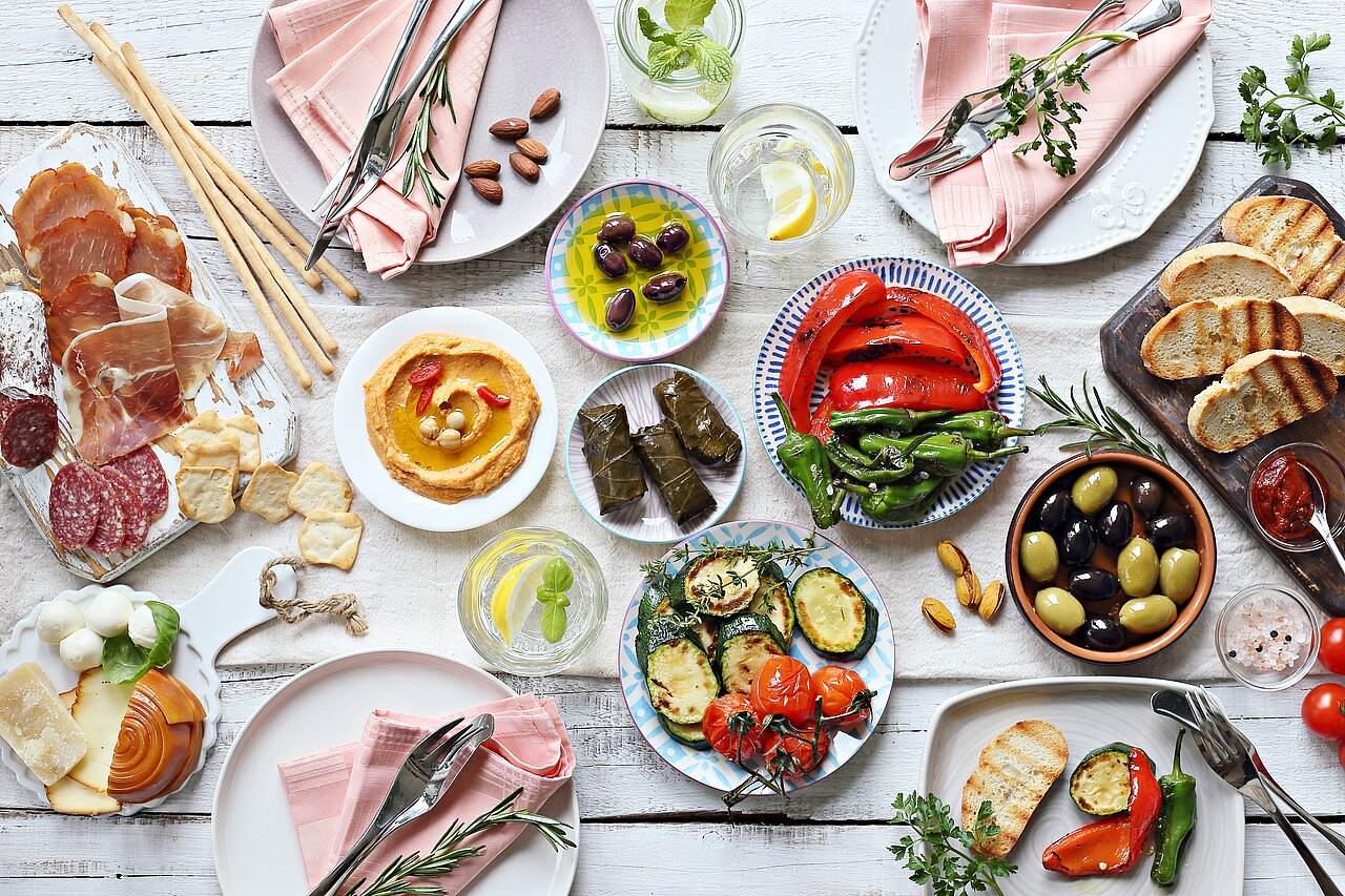 Mediterranean Appetizer Table Setting. Dinner table with a selection of antipasti: grilled vegetables, cold cuts, meat and cold cuts, ham, olives, cheese, hummus, and toasted bread.
